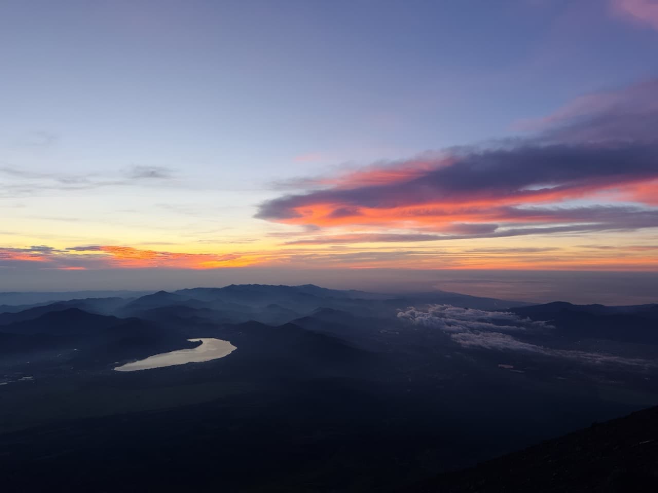 Dawn from a mountain summit: pink and orange clouds over layered blue ridgelines, with a lake and villages far below