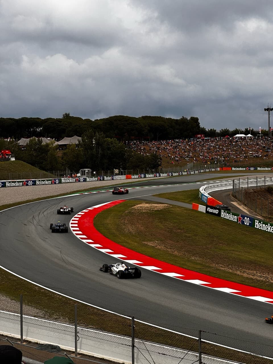 Open-wheel race cars sweeping through a red-and-white curbed corner at the Spanish Grand Prix as fans watch from a grassy hillside