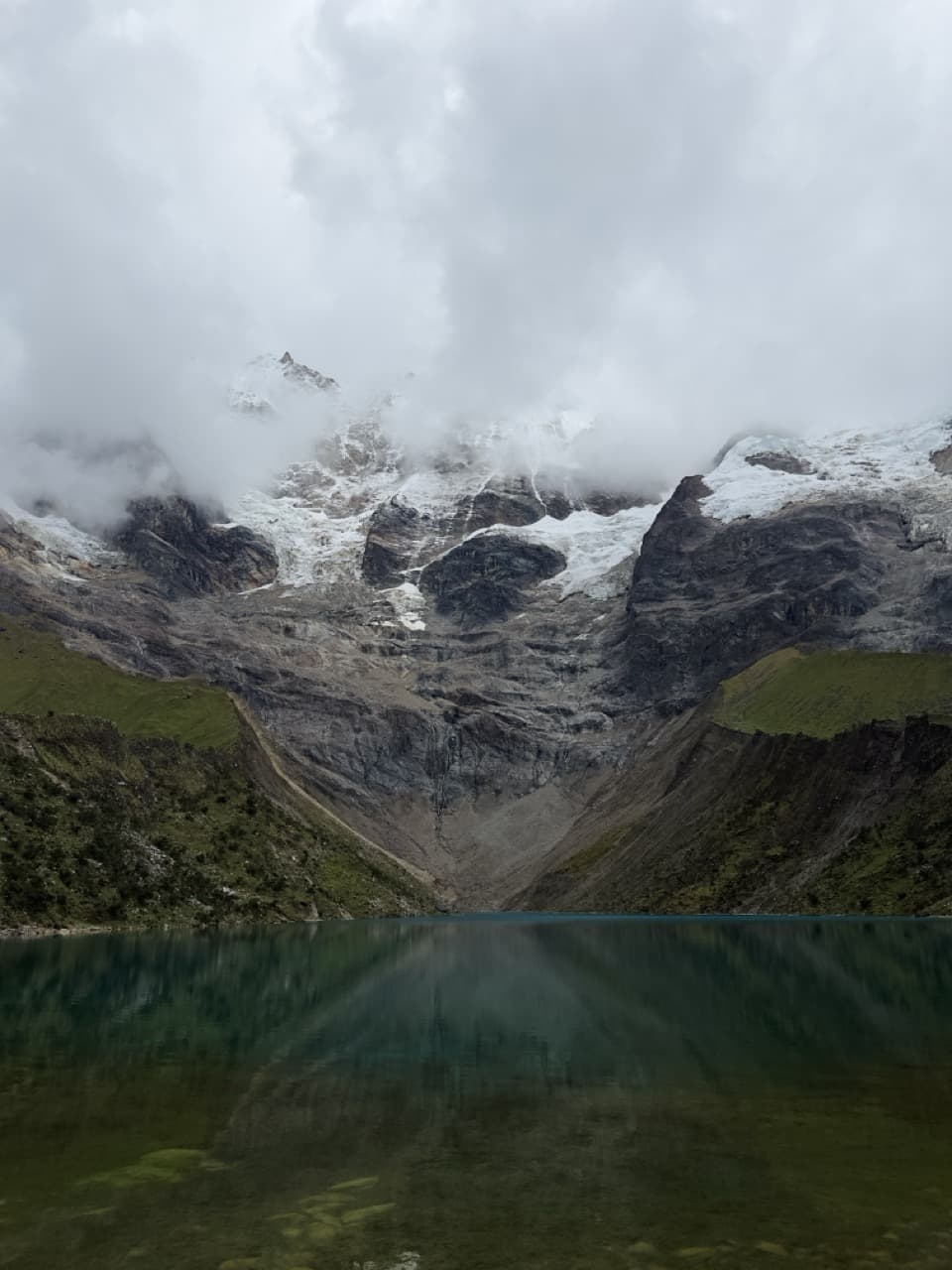 A still glacial lake reflecting green hillsides beneath a snow-covered peak shrouded in clouds