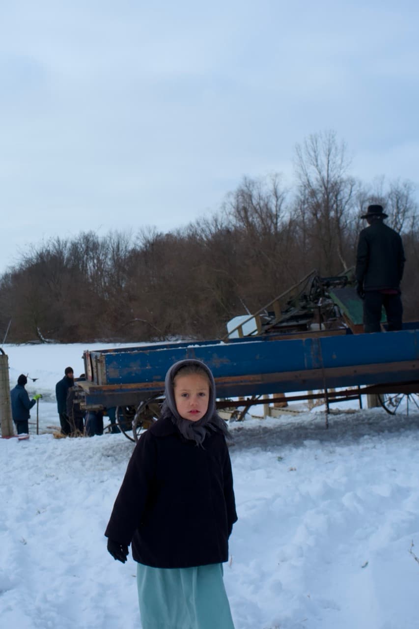 A young Amish girl in a headscarf and dark coat standing in the snow, with a blue wagon and ice harvesters working behind her